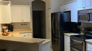 Una cocina con gabinetes blancos, un refrigerador negro, un microondas y una estufa. Hay un letrero en la pared que dice 'cocina, por favor'.