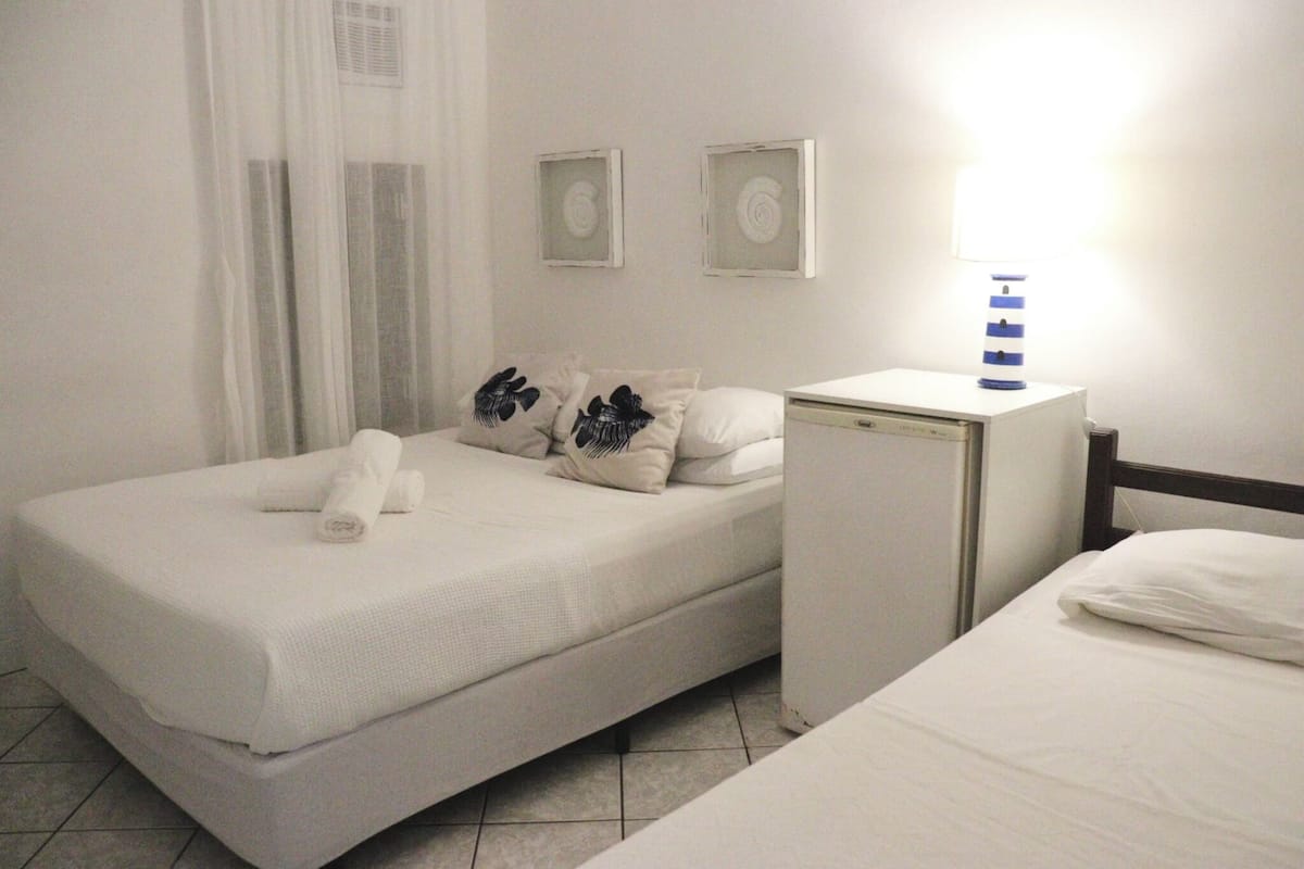 Een enkel bed met een witte matras en kussens, een klein nachtkastje met een lamp, en een wandairconditioner.