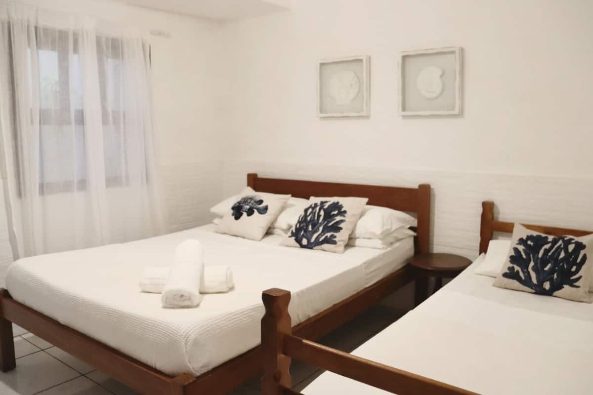 Een slaapkamer met een houten bed, witte beddengoed en decoratieve kussens. Er zijn twee nachtkastjes en een raam met dunne gordijnen.