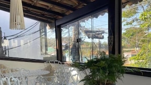 A covered outdoor seating area with white metal chairs and a table, a potted plant, and a view of trees and buildings through large windows. - Peixe Crew Rua das Pedras (Búzios)
