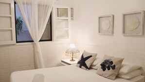 A bedroom with a bed, a nightstand with a lamp, a window with a view, and decorative shells on the wall. - Peixe Crew Rua das Pedras (Búzios)