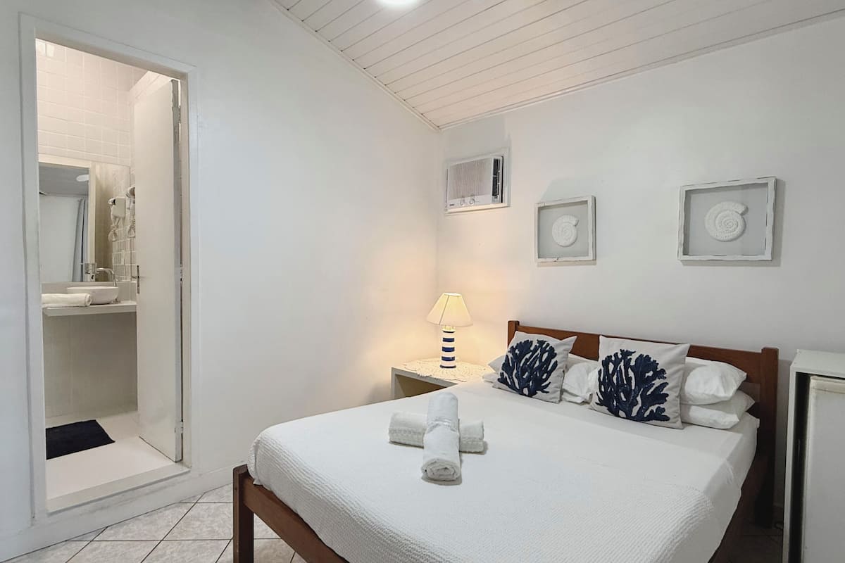 Een slaapkamer met een bed, nachtkastje, lamp en een badkamer door een geopende deur zichtbaar.
