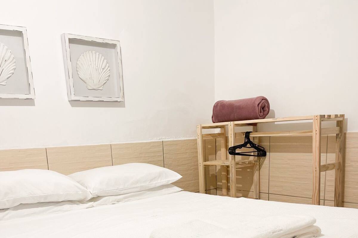 Een net opgemaakt bed met witte lakens, een houten hoofdbord en een houten nachtkastje met een roze handdoek en een zwarte hanger.