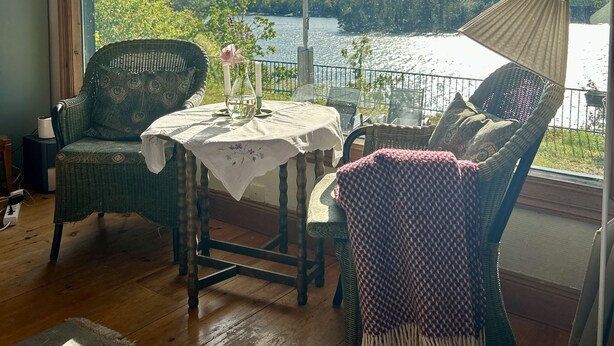 A cozy room with a view of a lake, featuring wicker furniture, a table with a vase and candles, and a large window with a view of trees and a fence.