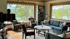 A cozy living room with a wooden ceiling, a fireplace, a bookshelf, a sofa, and a view of trees through large windows.