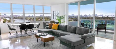 A spacious living room with a gray sectional sofa, a coffee table, and a dining area with a white table and black chairs. Large windows offer a view of a city skyline and a body of water.