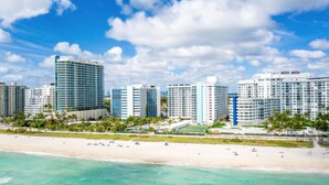 A beachfront hotel with multiple high-rise buildings, a sandy beach, and clear blue skies. - 2bd Beachfront Master Ste, Bay View w/balcony, Pool, nearby dining, beach access (Miami Beach)