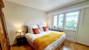 A bedroom with a bed, a nightstand, a lamp, and a window with a view of buildings. - New 2bedrooms in Center Terrace&Parking (Luxembourg City)