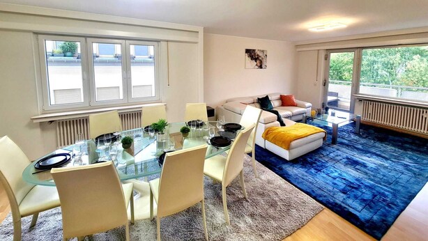 A modern living room with a glass dining table, white sofas, and a blue rug. - New 2bedrooms in Center Terrace&Parking (Luxembourg City)