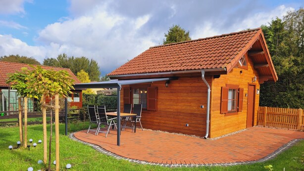 Et træhus med et tegltag, en overdækket terrasse med et bord og stole, og en indhegnet gård.
