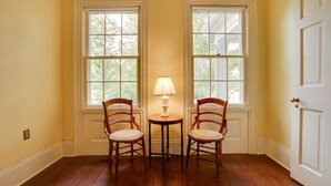 A room with wooden flooring, two windows with white frames, a table with a lamp, and two wooden chairs.