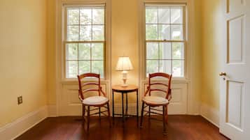 Une pièce avec un plancher en bois, deux fenêtres à cadres blancs, une table avec une lampe et deux chaises en bois.
