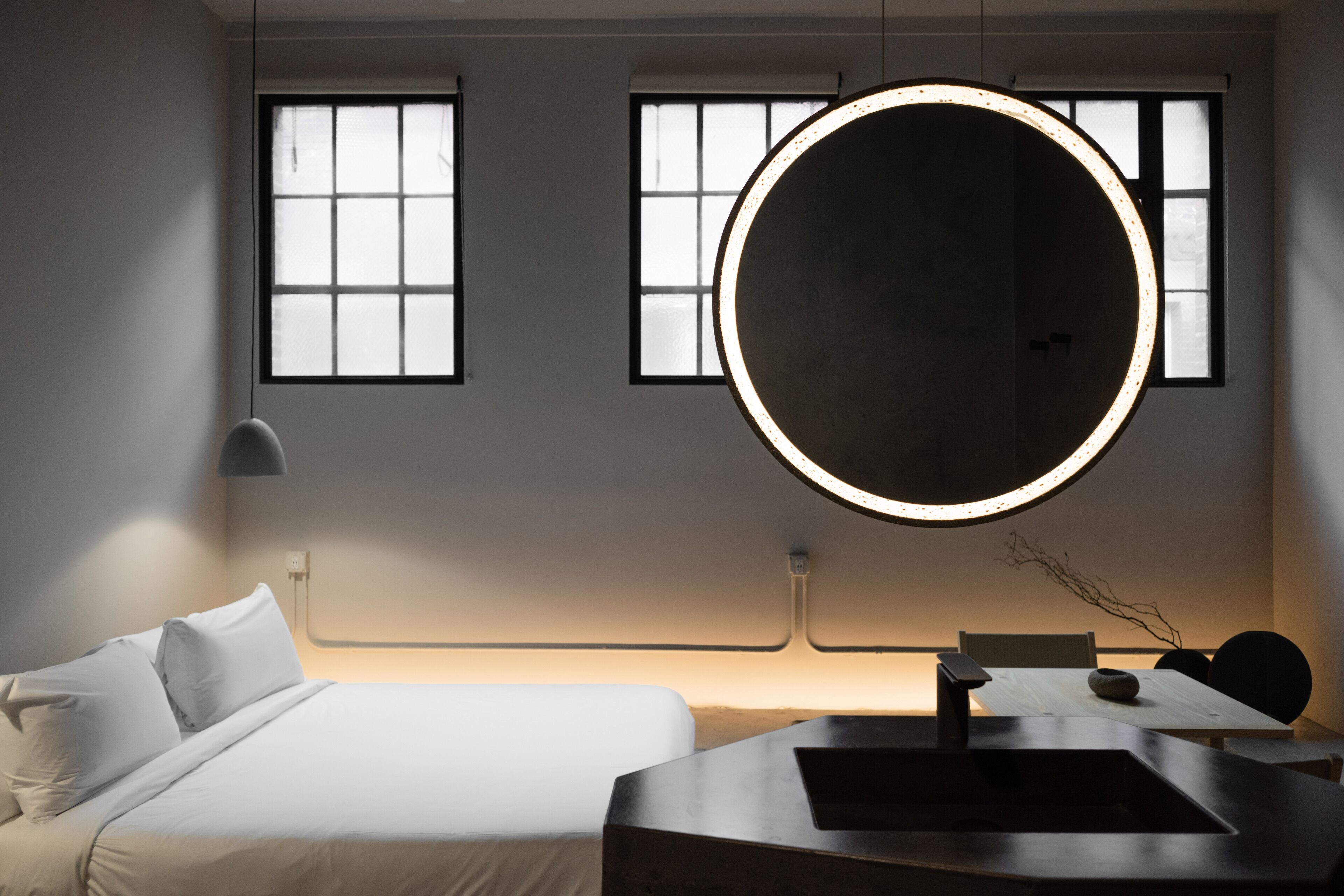 A modern hotel room with a bed, a round mirror, a table, and two windows with blinds.
