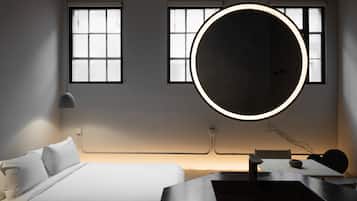A modern hotel room with a bed, a round mirror, a table, and two windows with blinds.