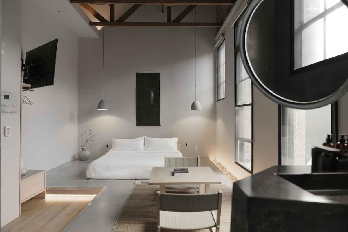 A modern bedroom with a bed, bedside table, and a small desk with a chair. There is a large window providing natural light and a view of the outside.