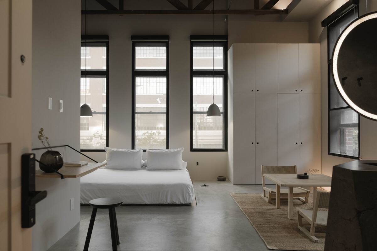 A modern bedroom with a bed, bedside table, and a desk with a chair. There are large windows and a wardrobe.