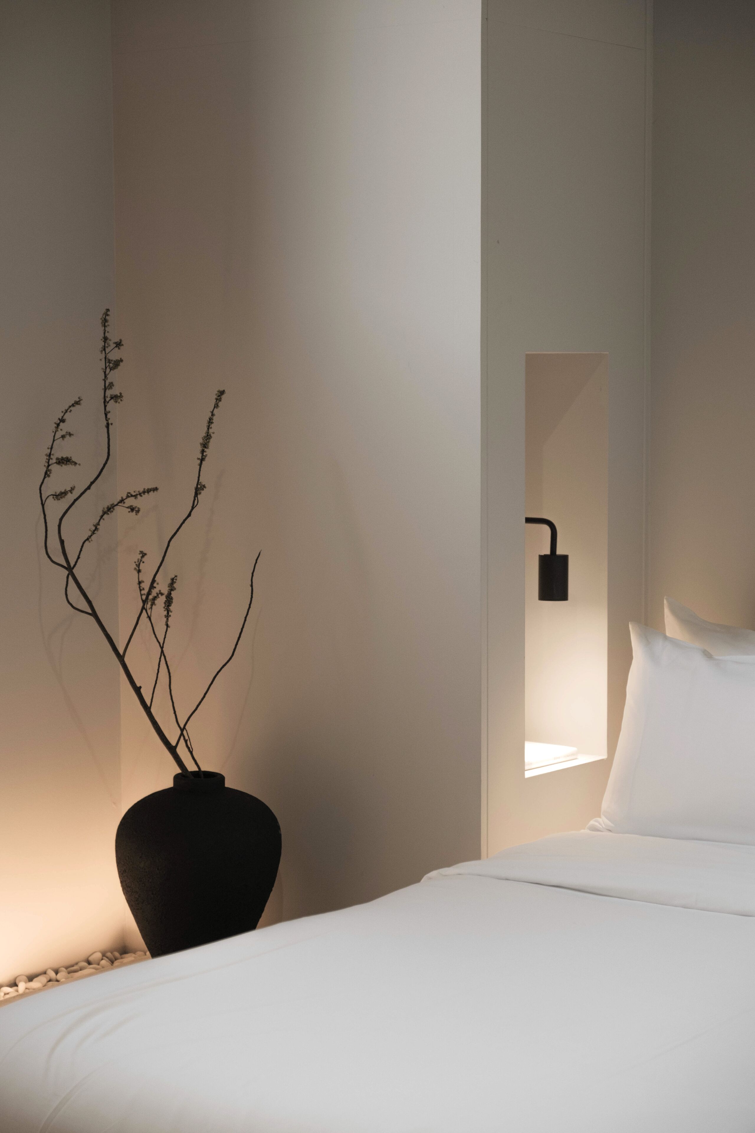A bedroom with a bed, a vase with branches, a wall-mounted light, and a mirror.