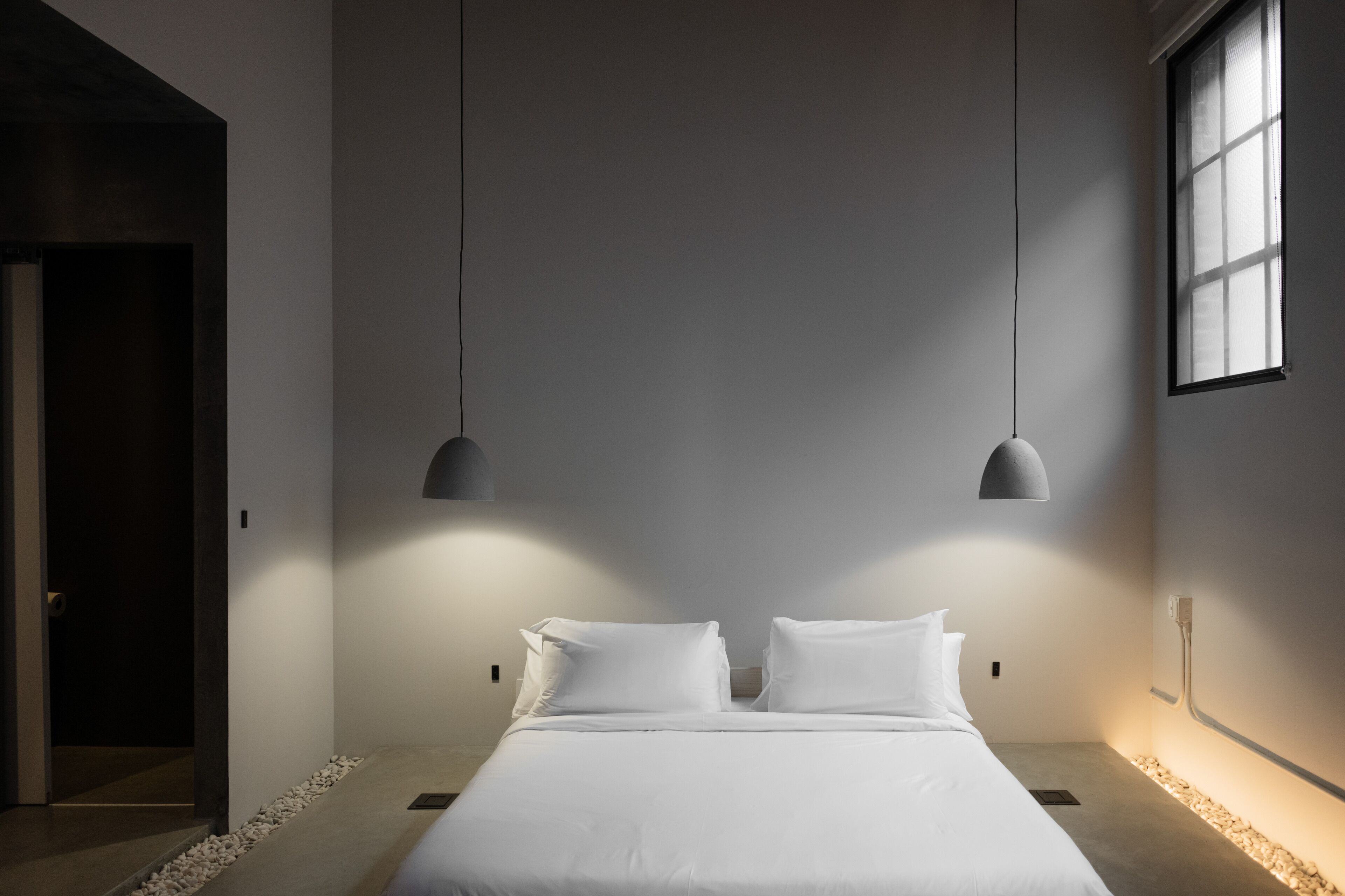 A minimalist bedroom with a large bed, two pendant lights, and a window on the right.