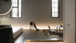 Quarto minimalista com uma cama, uma cadeira, uma mesinha e um espelho grande. Duas janelas proporcionam luz natural.