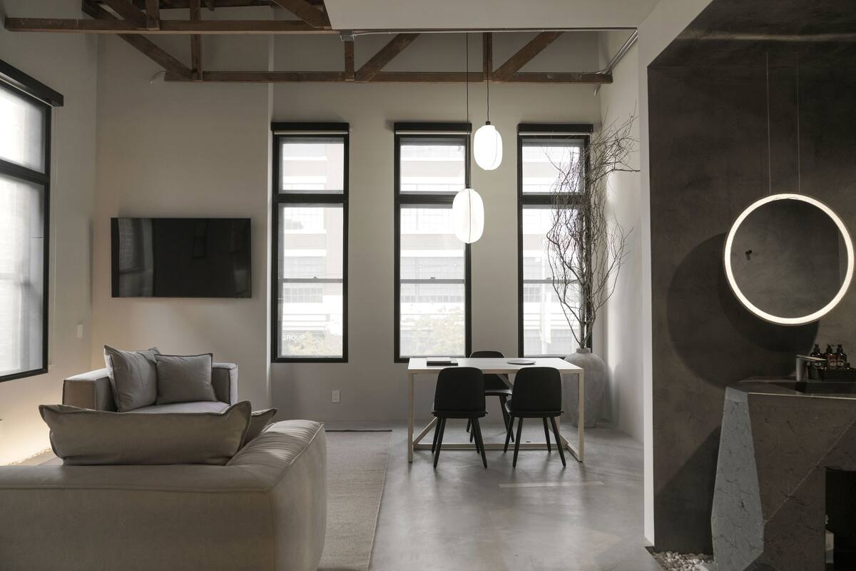 A modern living room with a sofa, a dining table with chairs, large windows, and a TV mounted on the wall.