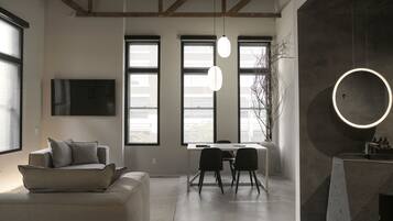 A modern living room with a sofa, a dining table with chairs, large windows, and a TV mounted on the wall.