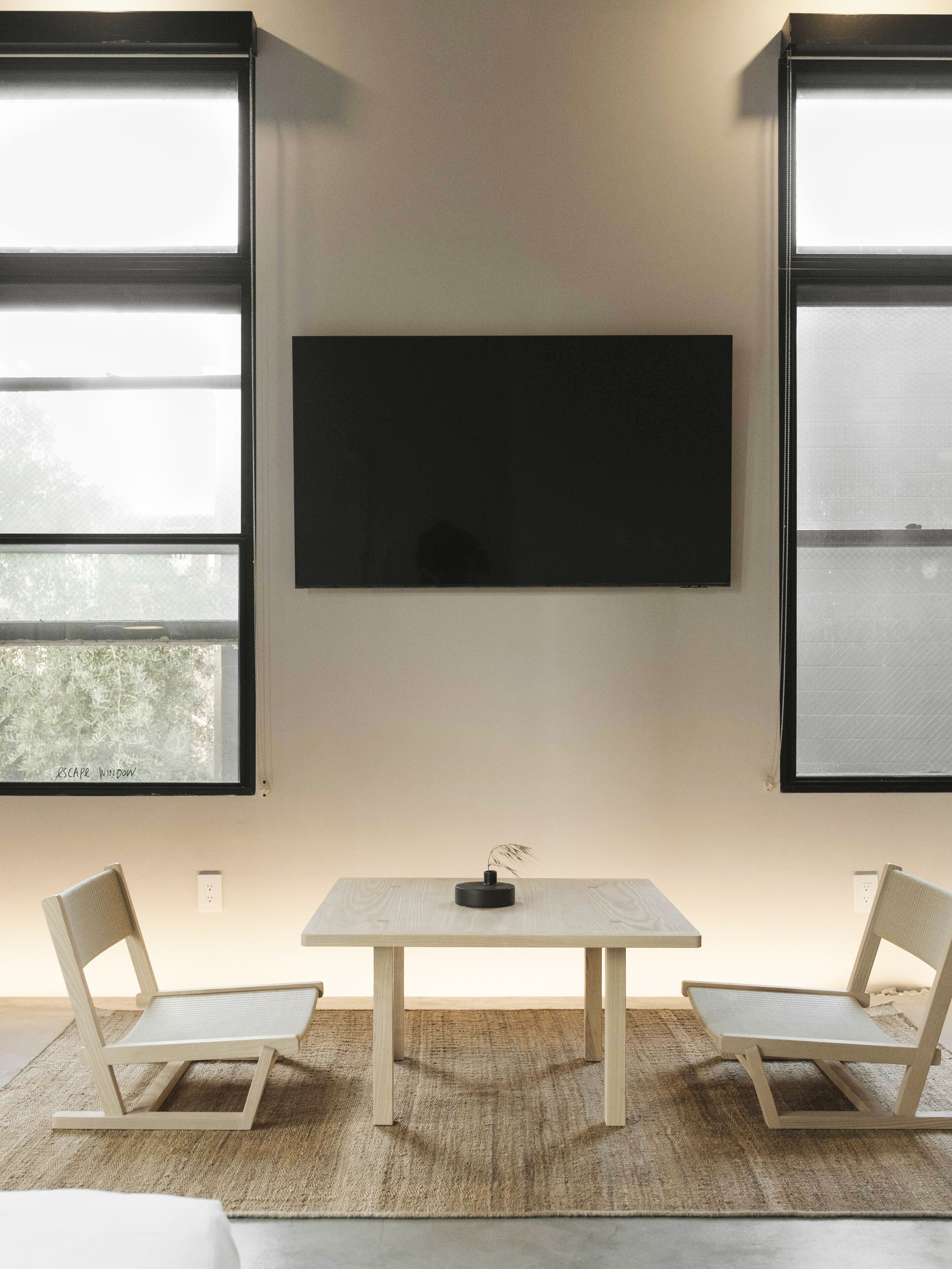 A minimalist room with a large flat-screen TV mounted on the wall, a small table with a vase, and two chairs.