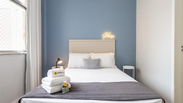 A neatly made bed with a grey blanket, a stack of white towels, and a small table with a lamp and toiletries.