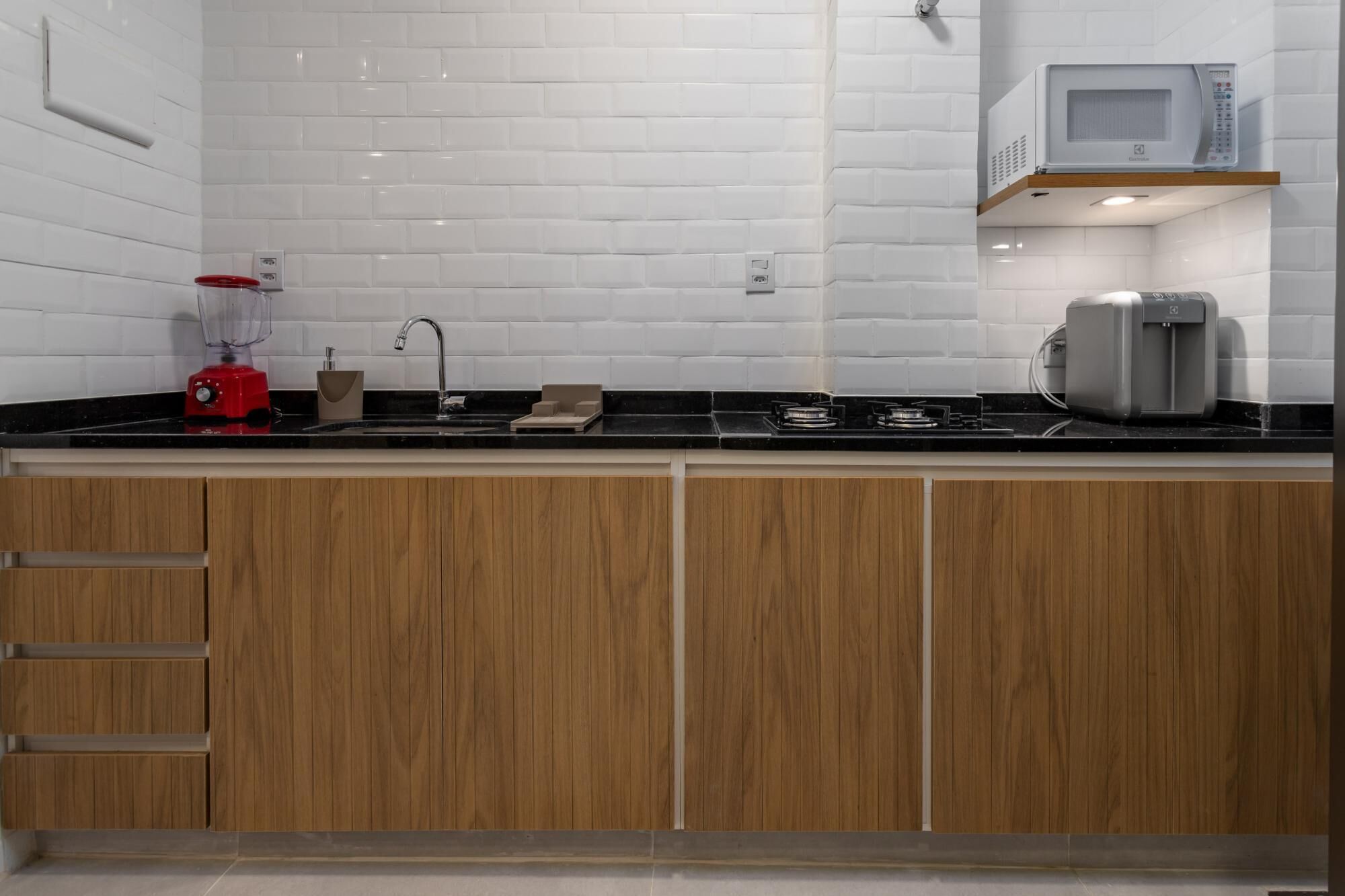 A modern kitchen with white subway tiles, a black countertop, wooden cabinets, a red blender, a microwave, and a coffee maker.