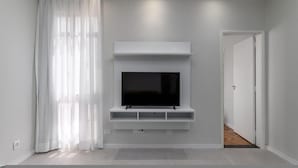 A modern living room with a flat-screen TV mounted on a white wall, a large window with sheer curtains, and a door leading to another room.