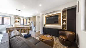 Ein modernes Wohnzimmer mit einem Ledersofa, einem runden Couchtisch und einem braunen Ledersessel. Ein großes Flachbildfernsehen ist an einer dunklen Holzvertäfelung an der Wand befestigt.