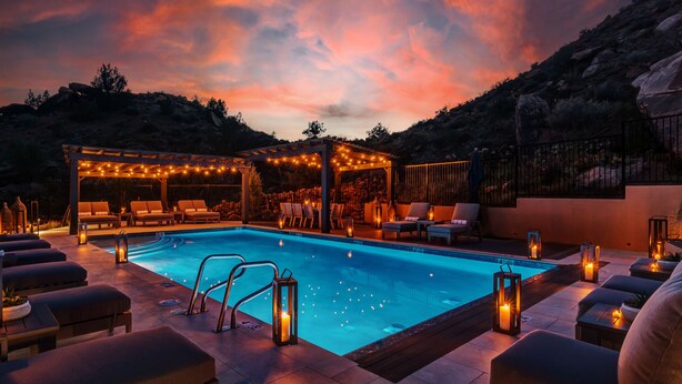 Un espace aménagé au bord de la piscine, avec des chaises longues, une pergola illuminée de guirlandes lumineuses et une vue sur les montagnes.