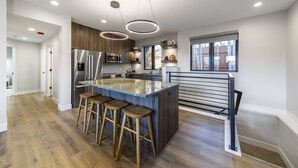 Cocina moderna con una isla central, taburetes de madera, electrodomésticos de acero inoxidable y un ventanal que proporciona luz natural.