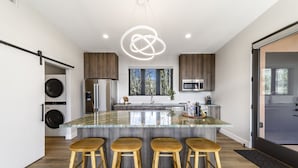 A modern kitchen with a central island, wooden stools, and a view of the outdoors through the window.