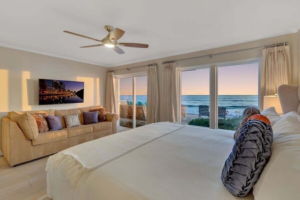 A hotel room with a large bed, a sofa, a ceiling fan, a TV, and a view of the ocean.