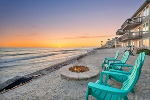 A beach scene with a fire pit and turquoise chairs, overlooking the ocean at sunset.