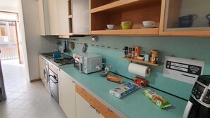 Cuina amb fogó de gas, microones, torradora i cafetera. Hi ha armaris sobre la barra i una prestatgeria amb plats.