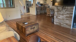 A spacious living room with a wooden floor, a vintage suitcase, and a leather couch. A kitchen with modern appliances and a dining area with wicker chairs is visible in the background. - 2 bed/ 2.5 bath Waterfront Condo with 2 pools and great fishing (Bokeelia)