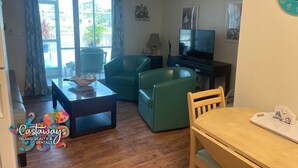 A living room with a TV, two green sofas, a wooden dining table, and a window with curtains. - Peaceful Island Living @ The Buccaneer Condo 1BR/1 BR with pool and dock (Saint James City)