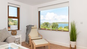 Ein Zimmer mit großem Fenster und Blick auf See und Grünfläche, ein Holzstuhl, ein weißer Tisch mit Glasplatte und ein Blumentopf.