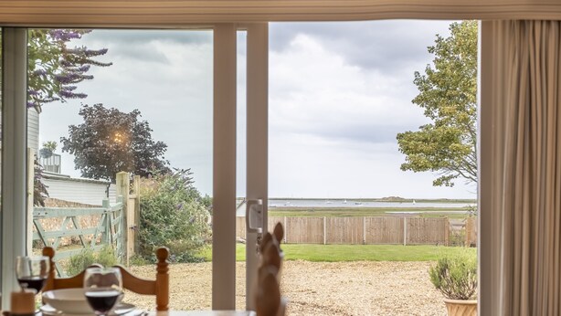 A dining area with a table set for two, overlooking a backyard with a wooden fence and a grassy lawn. - Blackhorse Cottage, Brancaster Staithe, Norfok (Brancaster Staithe)