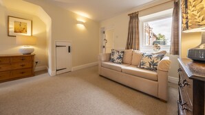 A cozy living room with a beige sofa, a wooden dresser, and a window with curtains. - Blackhorse Cottage, Brancaster Staithe, Norfok (Brancaster Staithe)