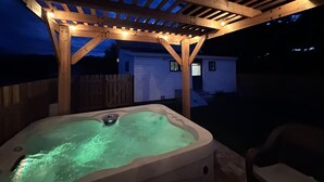 Ein Whirlpool unter einer Hollaube mit Lichterketten, umgeben von einer Holzterrasse und einem weißen Haus im Hintergrund.
