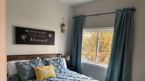 A bedroom with a bed, a bedside table, a lamp, a window with curtains, and a wall sign.