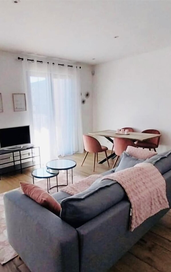 A modern living room with a grey sofa, a small round coffee table, a flat-screen TV on a black stand, a dining table with pink chairs, and a large window with sheer curtains. - Luxury 2 bedrooms with Parking&Terrace (Luxembourg City)