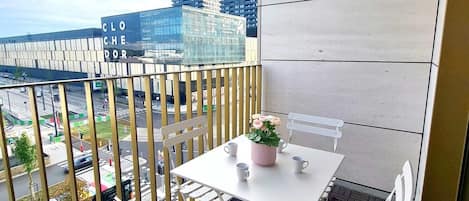 A balcony with a white table and chairs, a potted plant, and a view of a cityscape with buildings and a road.