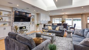 A spacious living room with a large flat-screen TV, grey sofas, a fireplace, and a dining area with natural light from large windows. - Fletcher Hills luxury, Sunset View, 4 BR, Saltwater Pool and Spa (El Cajon)
