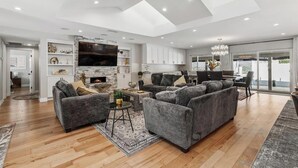 A spacious living room with a fireplace, grey sofas, a TV, and a dining area with a view. - Fletcher Hills luxury, Sunset View, 4 BR, Saltwater Pool and Spa (El Cajon)