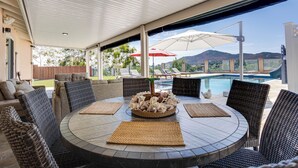 A covered outdoor seating area with wicker furniture, a round table with placemats, and a pool in the background. - Fletcher Hills luxury, Sunset View, 4 BR, Saltwater Pool and Spa (El Cajon)