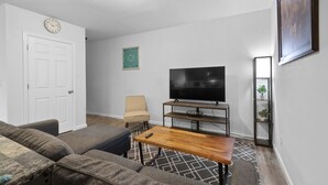 A living room with a sofa, a coffee table, a TV on a stand, a door, and a wall clock. - Modern Studio Apartment in the heart of Festus (Festus)
