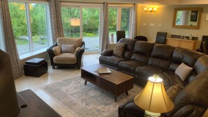 A spacious living room with a leather sofa, armchair, and ottoman, a coffee table, and a lamp. Large windows offer a view of greenery outside. - Viceroy home at Soames Hill (Gibsons)